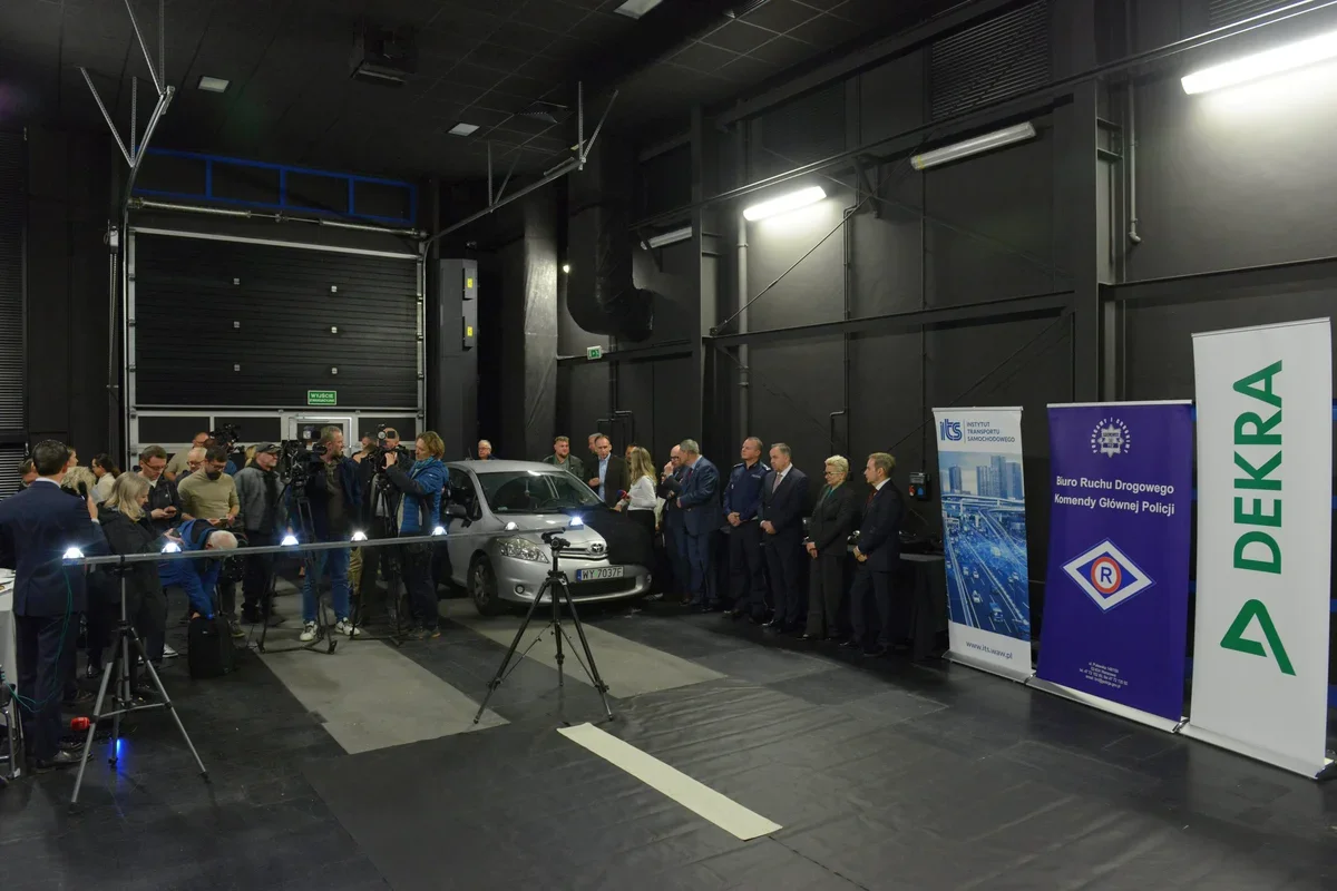 W hali przemysłowej odbywa się konferencja prasowa z udziałem przedstawicieli instytucji drogowych i DEKRA, obok stoi srebrny samochód osobowy.