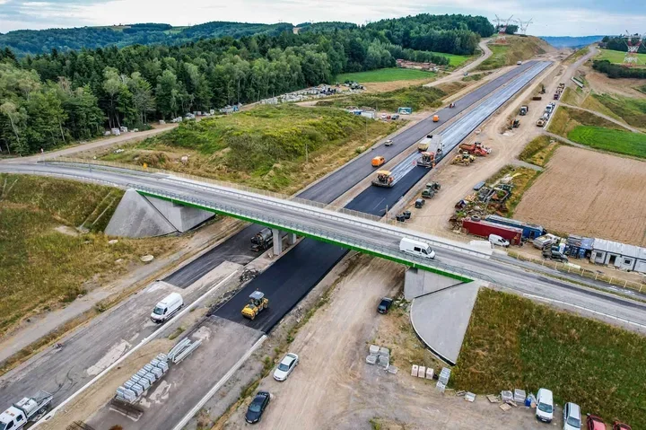 Robotnicy i maszyny budowlane układają asfalt na budowanej autostradzie, nad którą przeprowadzono nowy wiadukt drogowy w terenie zalesionym i rolniczym.