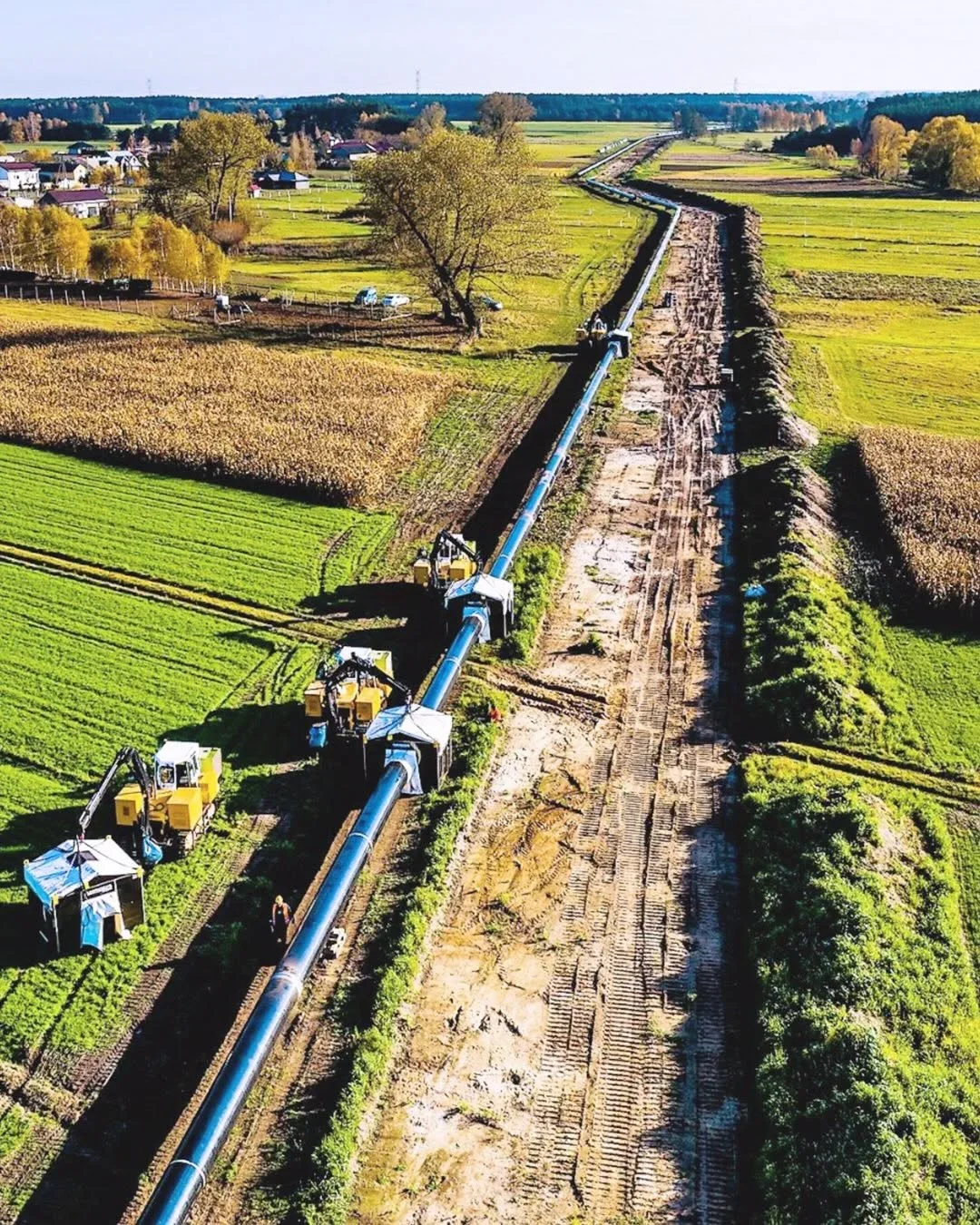 Robotnicy i maszyny budowlane układają dużą rurę pipeline'u na wykopanym wąskie w polach uprawnych.