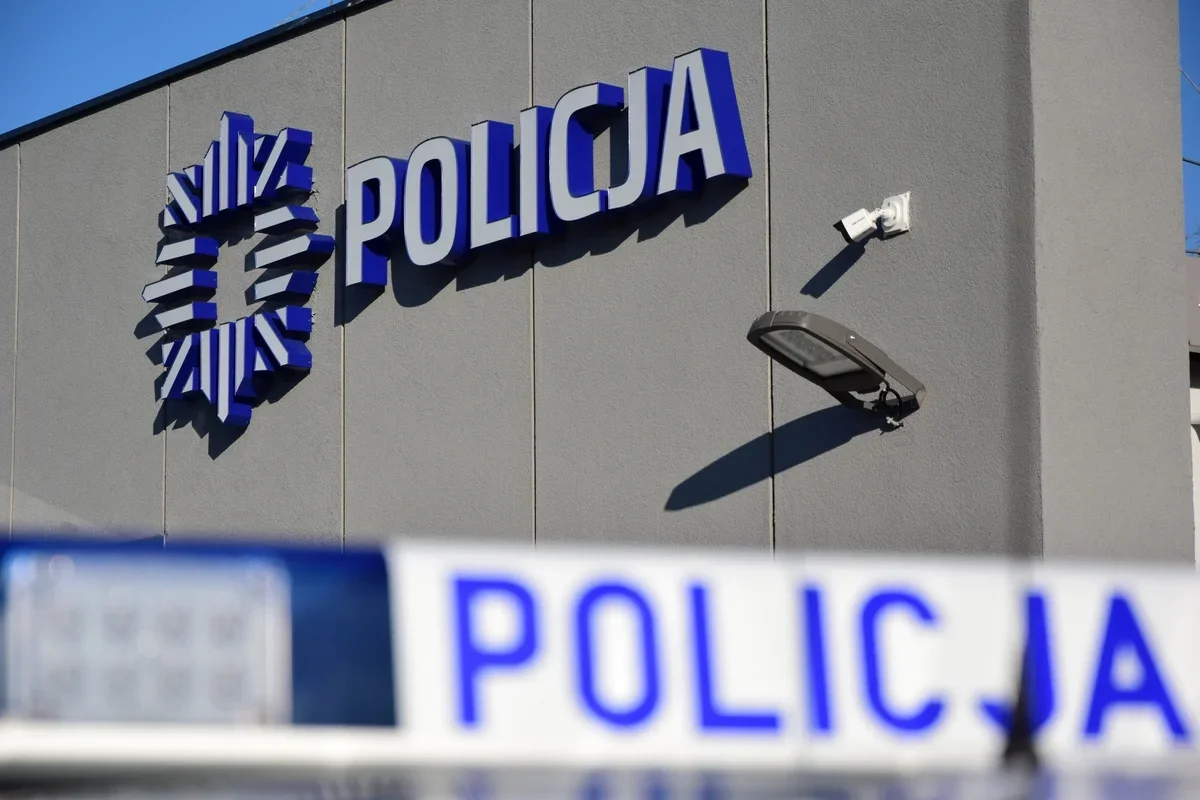 Fragment elewacji budynku z napisem i emblematem polskiej policji oraz policyjna taśma ostrzegawcza na pierwszym planie.