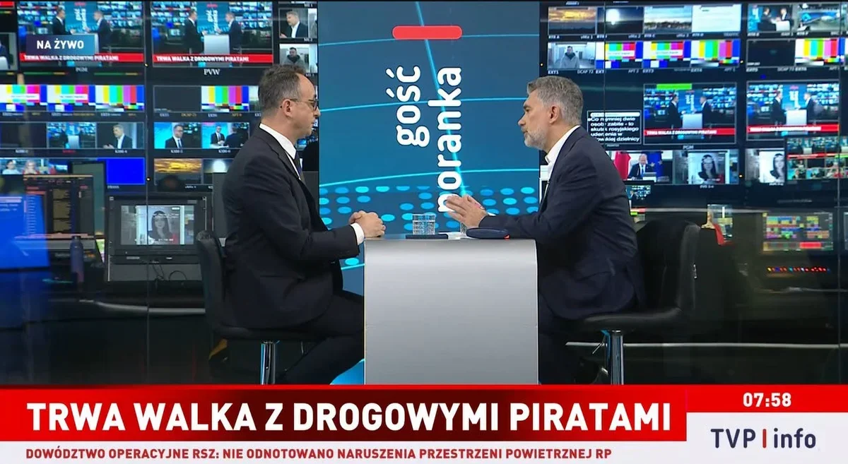 W studiu telewizyjnym TVP Info dwaj mężczyźni w garniturach prowadzą rozmowę przy stole, w tle ekrany monitoringu wizji.