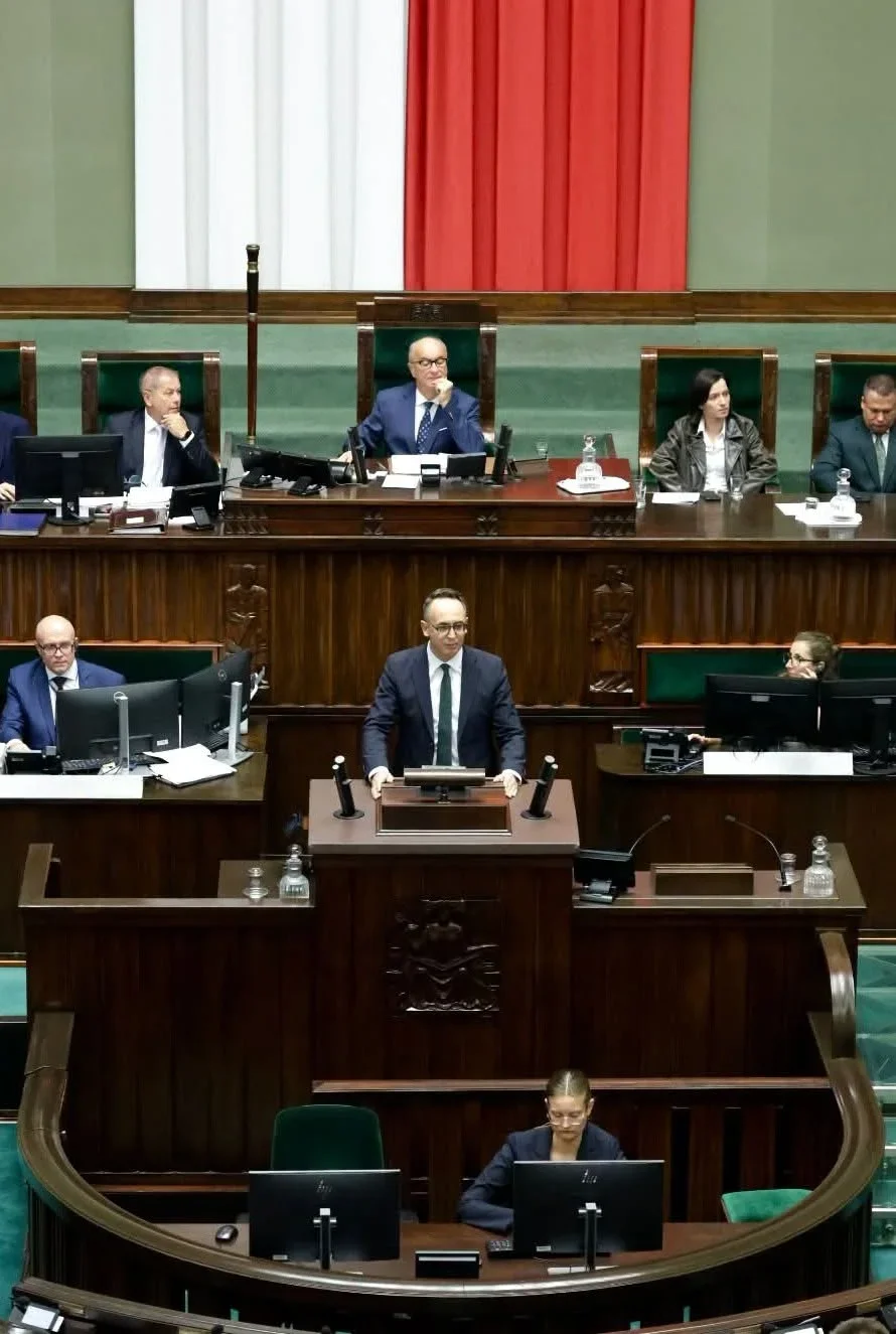 Mężczyzna w garniturze przemawia z mównicy w sali plenarnej parlamentu, za nim siedzą posłowie i przewodniczący obrad.