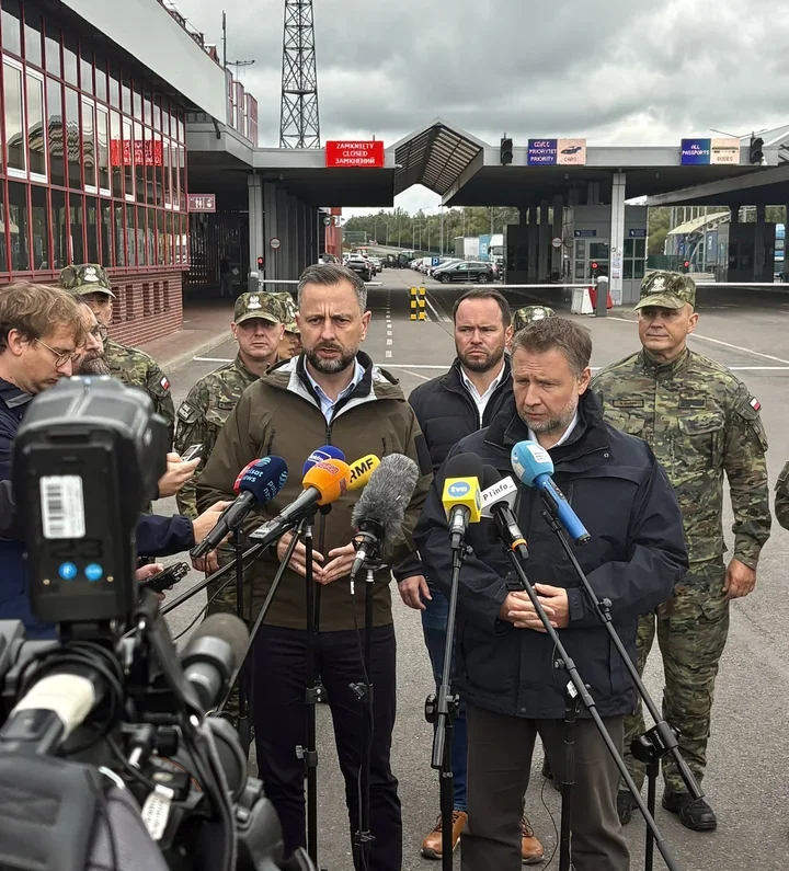 Dwaj mężczyźni w kurtkach stoją przed mikrofonami mediów na przejściu granicznym, za nimi żołnierze w mundurach i budynki z napisami wskazującymi zamkniętą drog