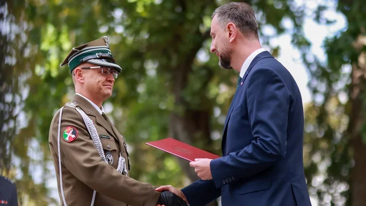 Mężczyzna w garniturze wręcza czerwoną teczkę oficerowi w brązowym mundurze podczas uroczystości na zewnątrz.