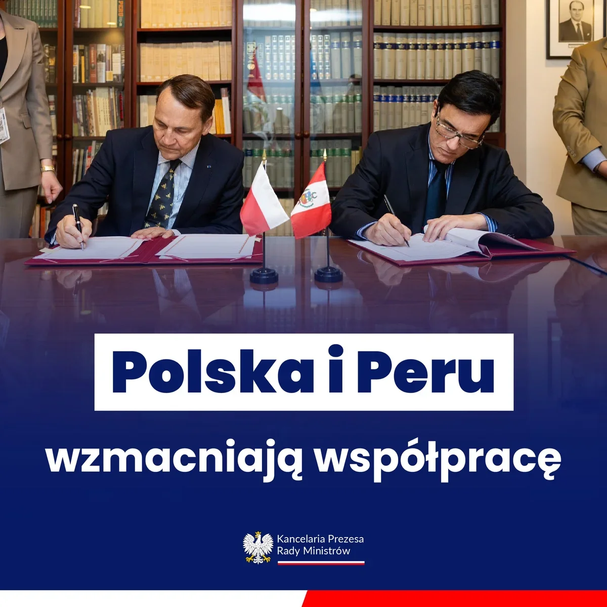 Dwaj mężczyźni w garniturach podpisują dokumenty przy stole, przy którym stoją małe flagi Polski i Peru, w pomieszczeniu z biblioteczkami.