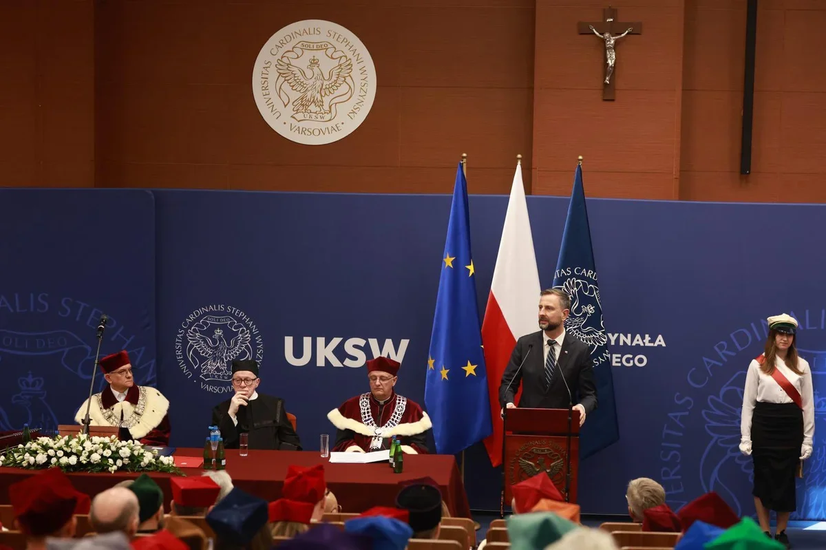 Mężczyzna w garniturze przemawia z pulpitu podczas uroczystej akademii w auli Uniwersytetu Kardynała Stefana Wyszyńskiego w Warszawie, przy stole siedzą trzej m