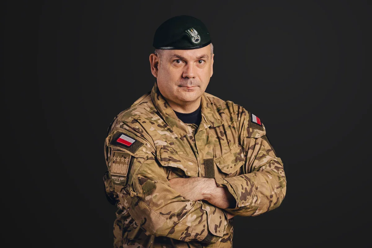 Mężczyzna w polskim mundurze wojskowym z beretem stoi z założonymi rękami na ciemnym tle.