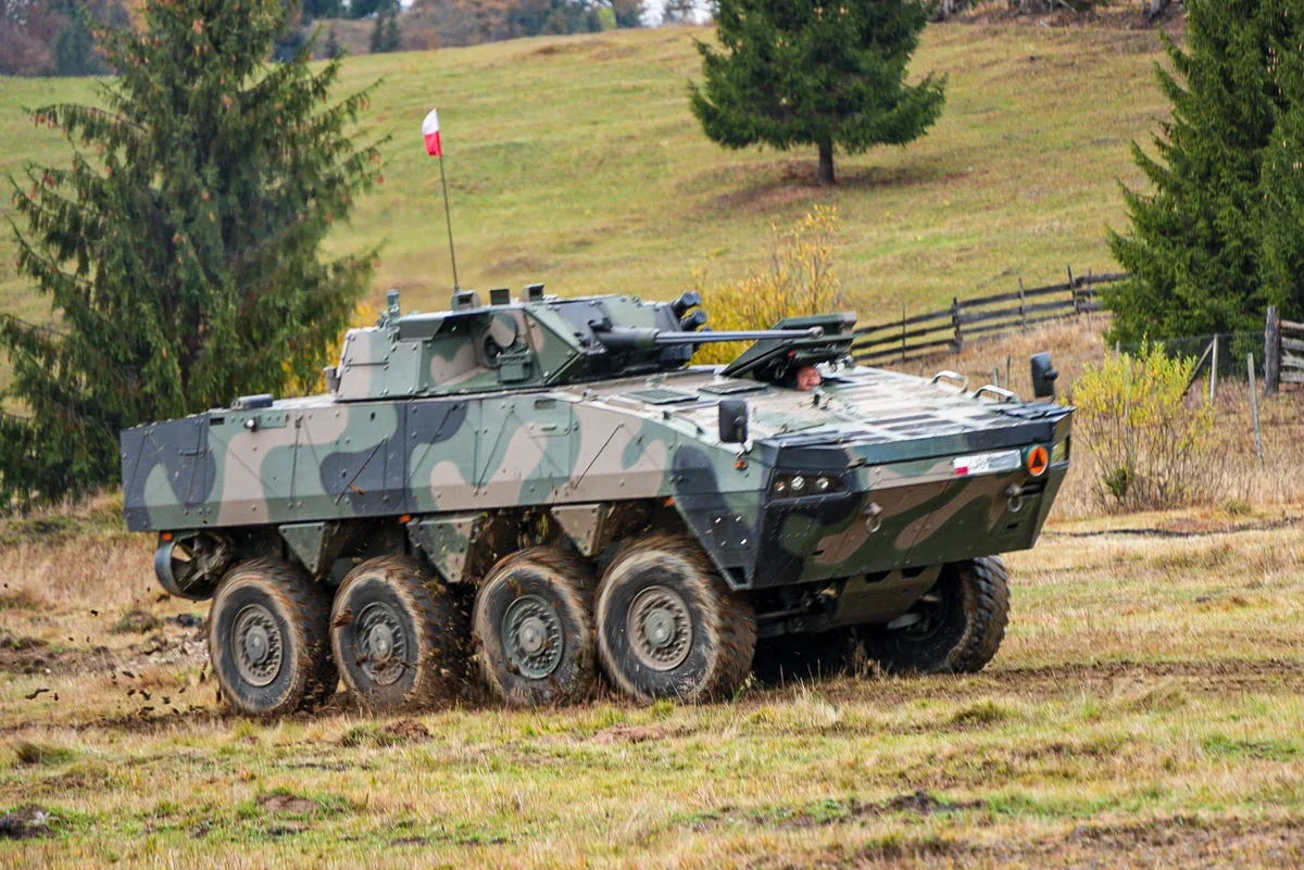 Wojskowy kołowy transporter opancerzony z polską flagą porusza się po polnej drodze w pagórkowatym terenie.