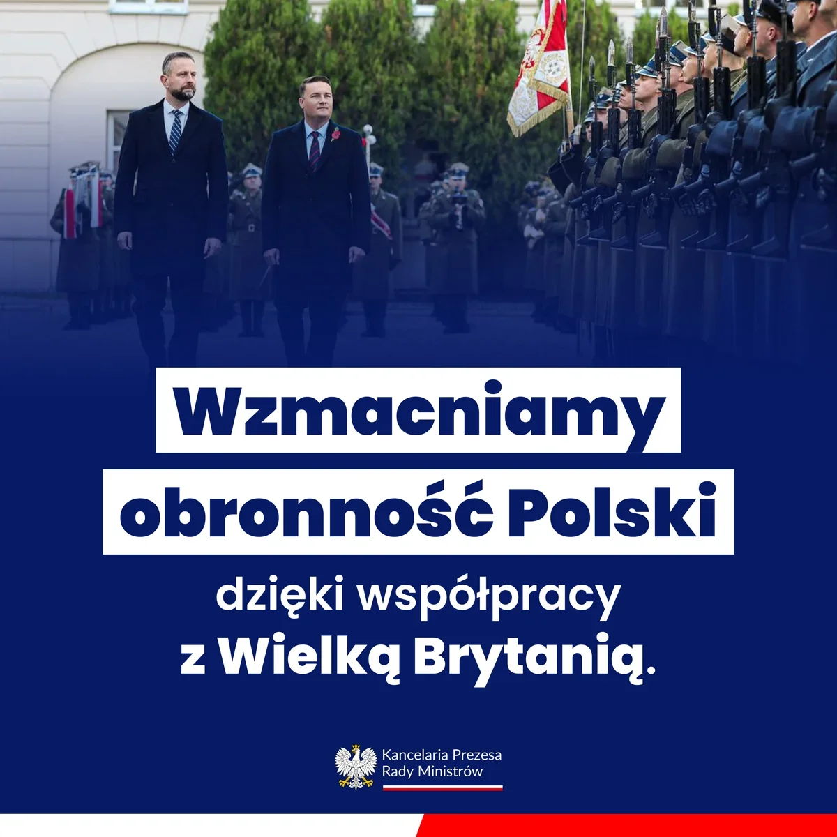Dwaj mężczyźni w ciemnych płaszczach przechodzą przed frontem honorowym podczas uroczystej defilady wojskowej.