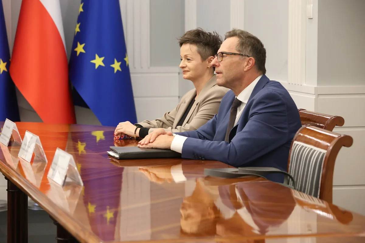 Dwie osoby siedzą przy lśniącym stole konferencyjnym z flagami Polski i Unii Europejskiej w tle.