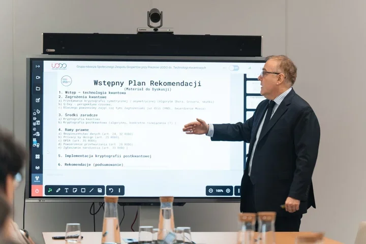 Mężczyzna w garniturze prezentuje slajd z planem rekomendacji dotyczących technologii kwantowych na dużym ekranie interaktywnym w sali konferencyjnej.