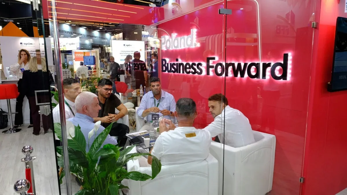 Grupa mężczyzn prowadzi rozmowę biznesową w szklanym boxie targowym z czerwoną ścianą z napisem „Poland. Business Forward” podczas wystawy branżowej.