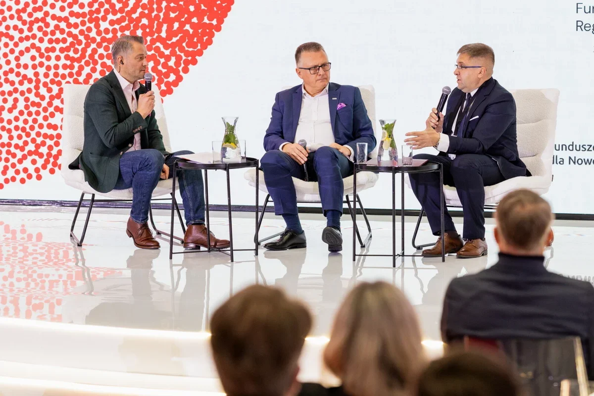 Trzech mężczyzn w garniturach uczestniczy w dyskusji panelowej na scenie przed publicznością.