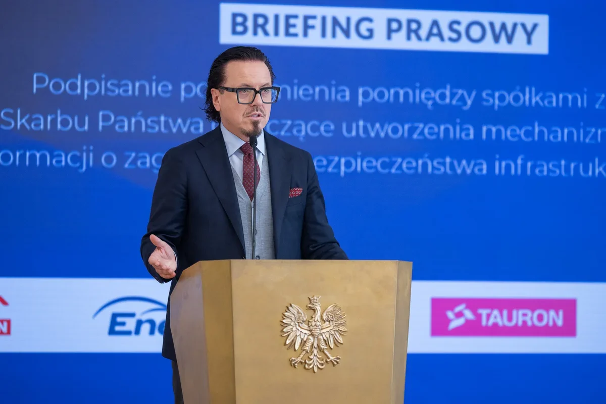 Mężczyzna w garniturze przemawia z brązowego pulpitu z orłem podczas briefingu prasowego, w tle logotypy spółek energetycznych i napis „BRIEFING PRASOWY”.