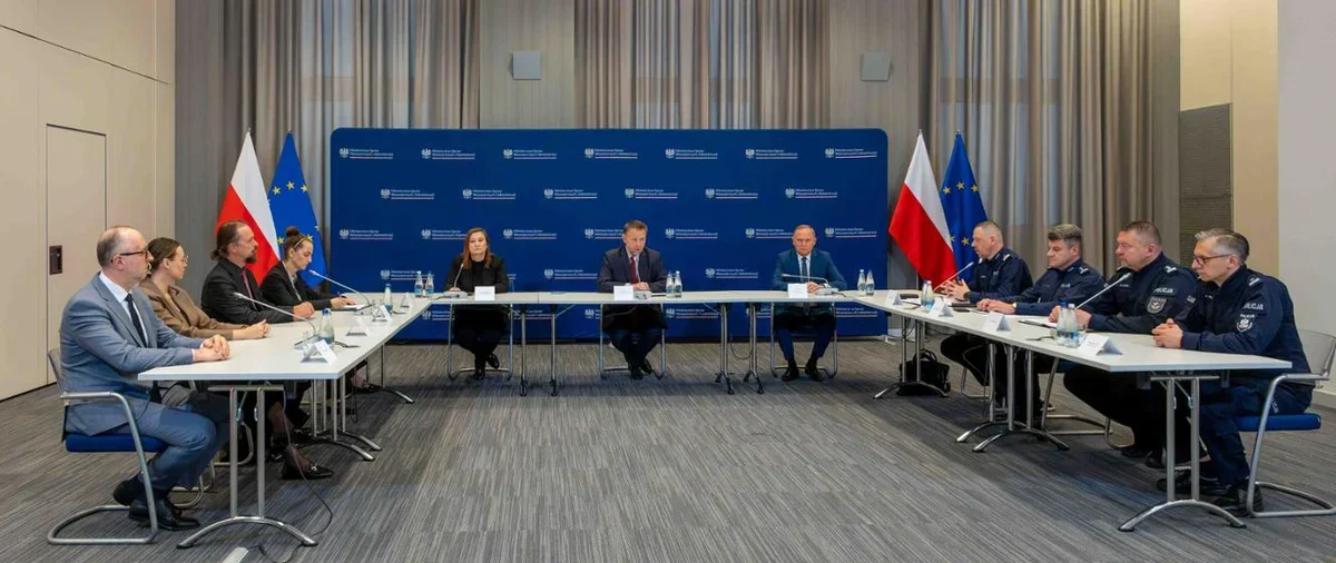 Spotkanie przy stole konferencyjnym w sali z flagami Polski i UE, z udziałem osób cywilnych i funkcjonariuszy policji.