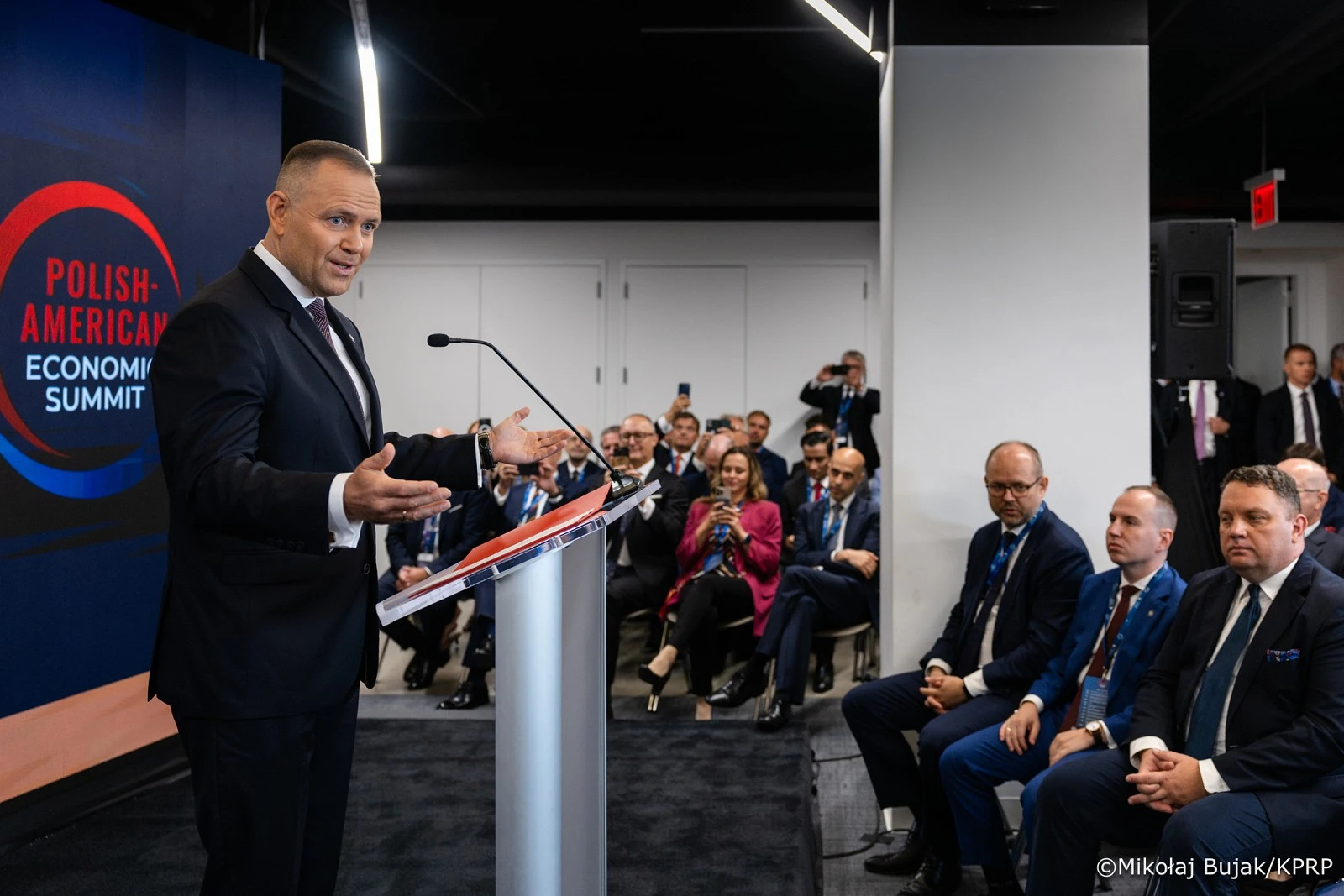 Mężczyzna w garniturze przemawia z mównicy podczas Polish-American Economic Summit, a przed nim siedzi grupa słuchaczy.