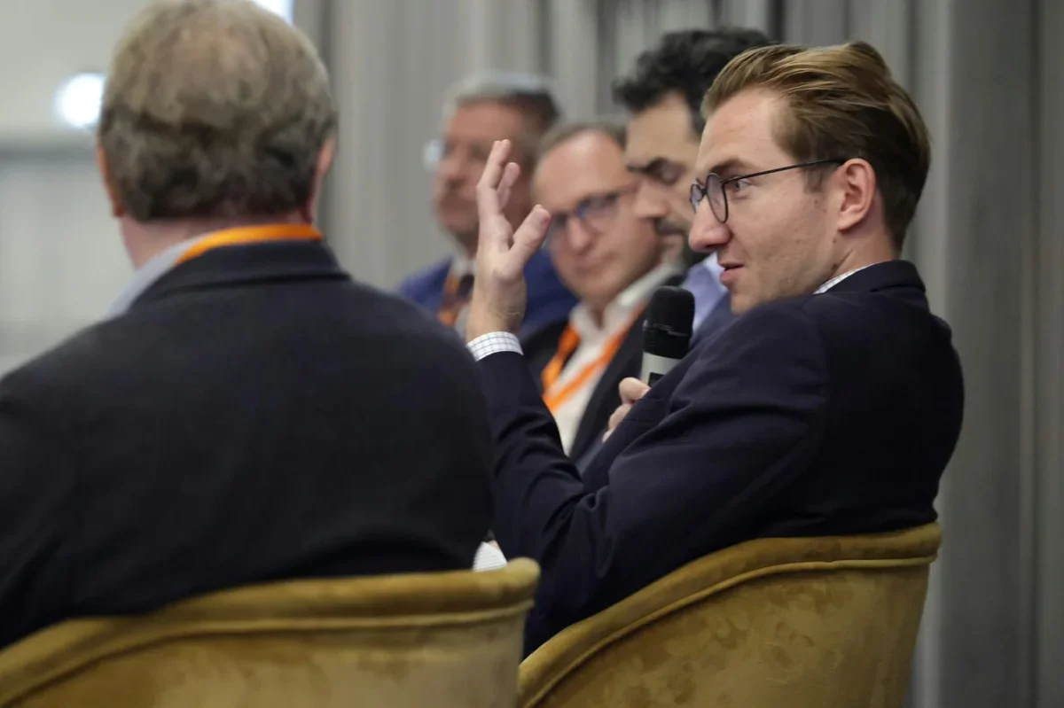 Mężczyzna w garniturze z mikrofonem gestykuluje podczas panelowej dyskusji w sali konferencyjnej.