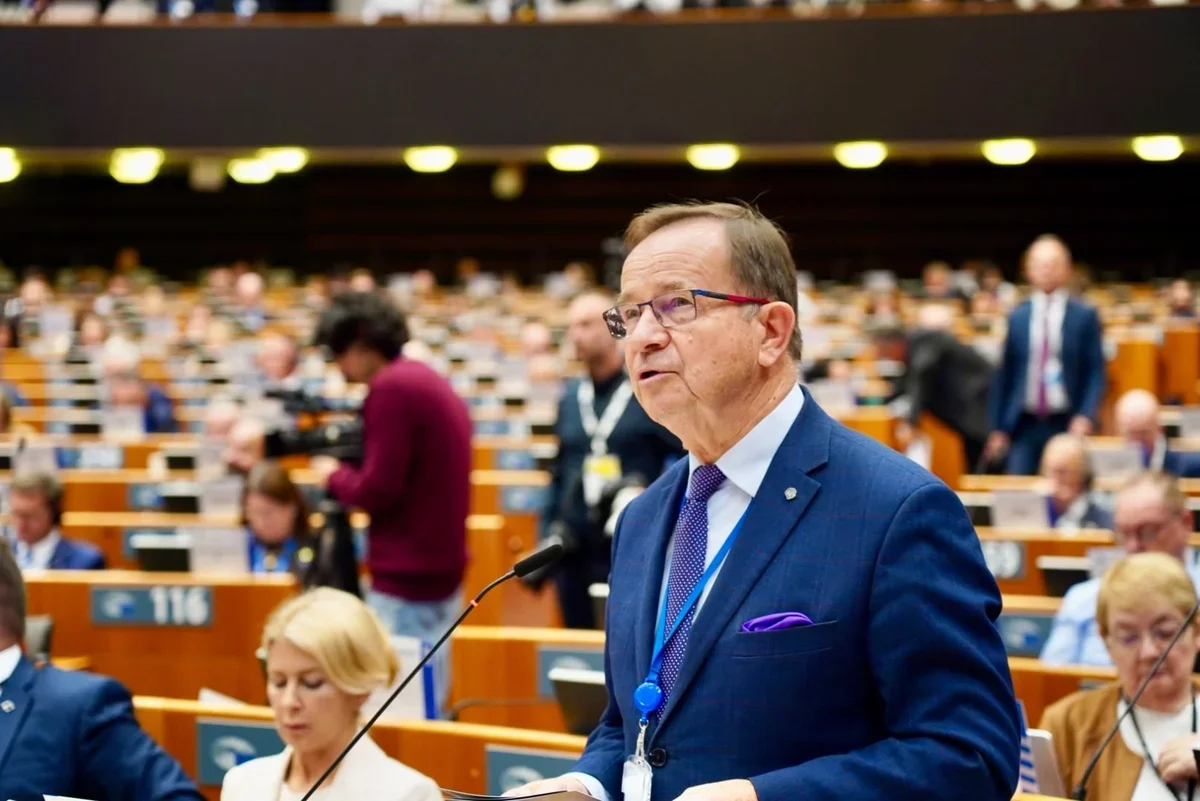 Mężczyzna w niebieskim garniturze przemawia z pulpitu w sali plenarnej parlamentu, w tle posłowie i dziennikarze.