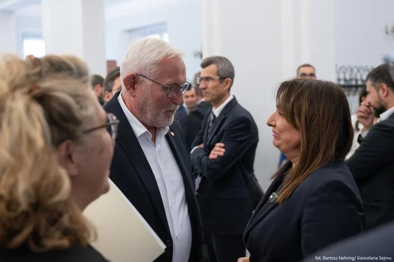 Mężczyzna z siwymi włosami w garniturze rozmawia z kobietą w ciemnym żakiecie podczas oficjalnego spotkania w białym wnętrzu.