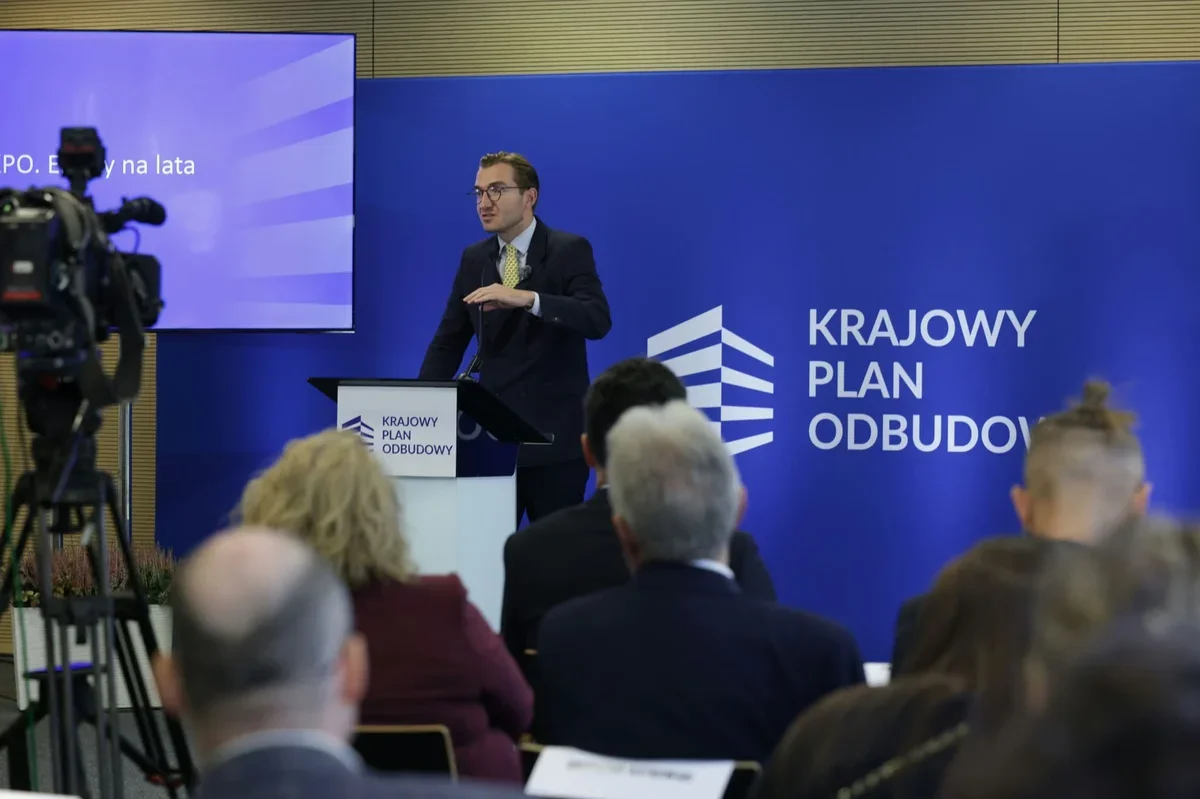 Mężczyzna w garniturze przemawia z mównicy podczas konferencji dotyczącej Krajowego Planu Odbudowy, w sali z publicznością i kamerą.