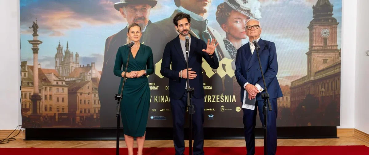 Trzy osoby stoją na czerwonym dywanie przy mikrofonach podczas konferencji prasowej przed plakatem filmowym.