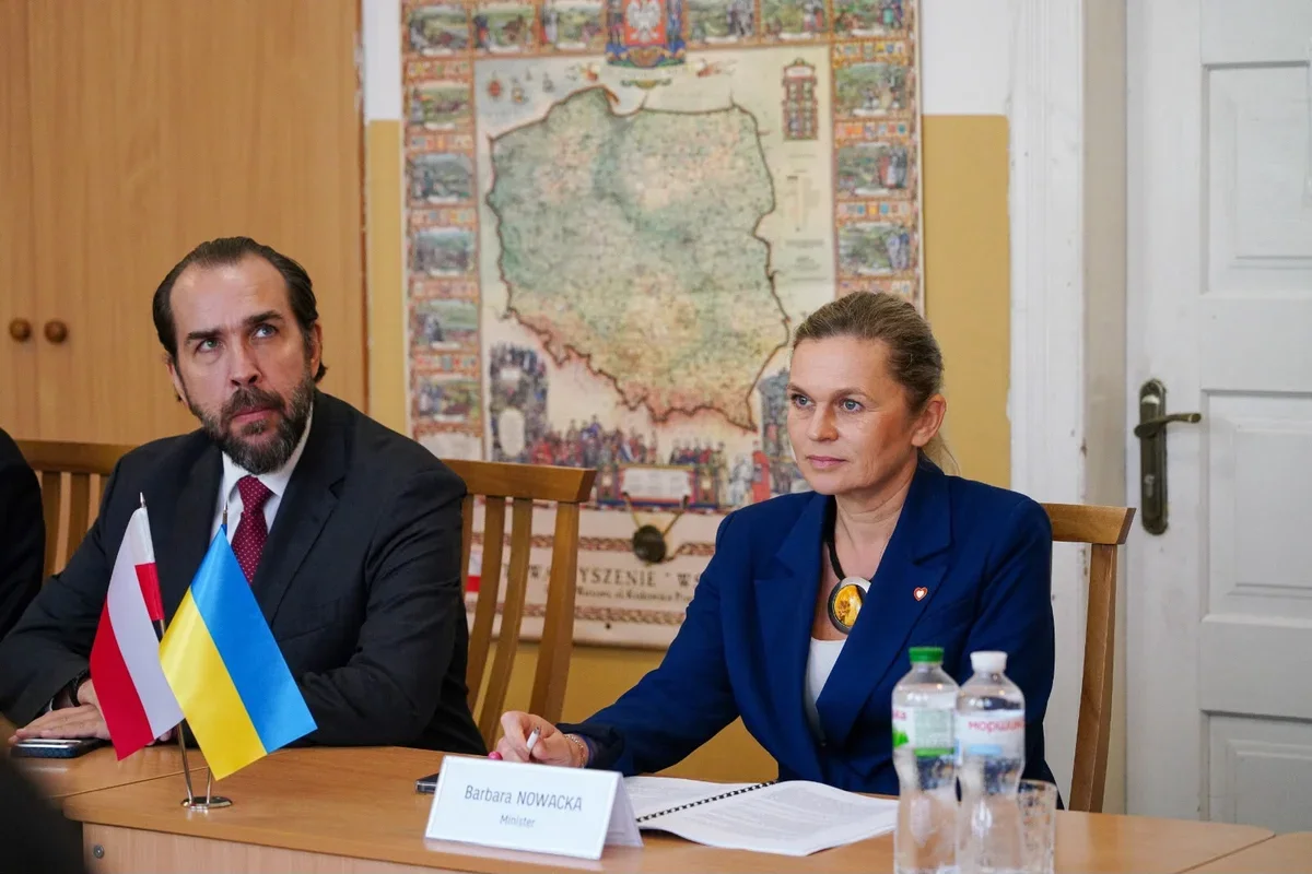 Dwie osoby siedzą przy drewnianym stole podczas spotkania formalnego, na którym stoją flagi Polski i Ukrainy oraz tabliczka z nazwiskiem Barbara NOWACKA.