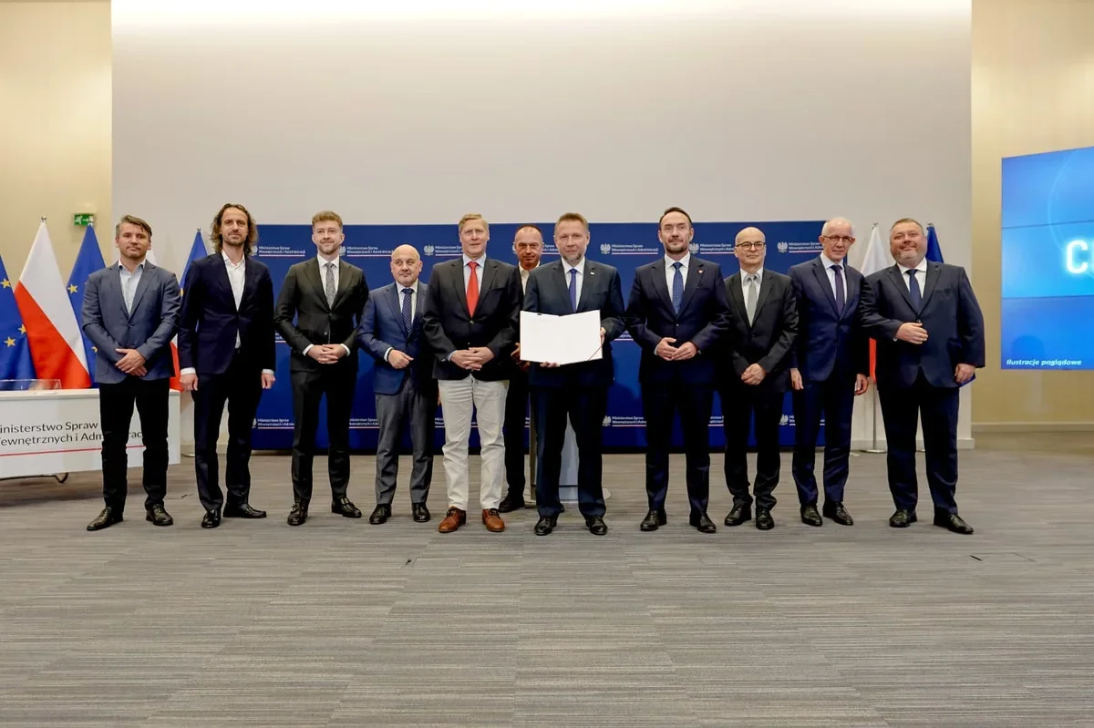 Grupa mężczyzn w garniturach pozuje do zdjęcia podczas oficjalnej uroczystości w siedzibie Ministerstwa Spraw Wewnętrznych i Administracji, jeden z nich trzyma