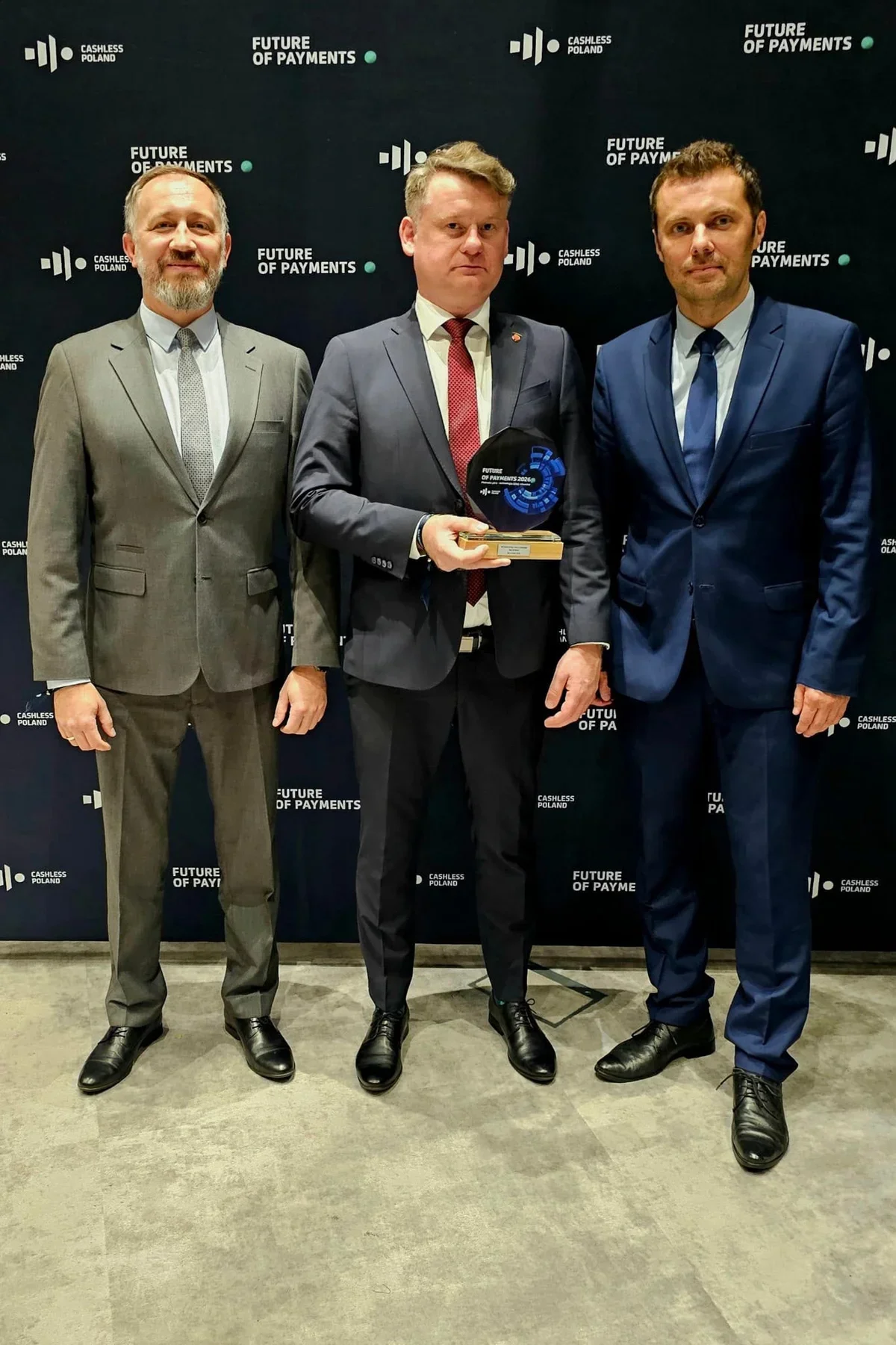 Trzej mężczyźni w garniturach pozują podczas gali Future of Payments organizowanej przez Cashless Poland, a mężczyzna pośrodku trzyma statuetkę.
