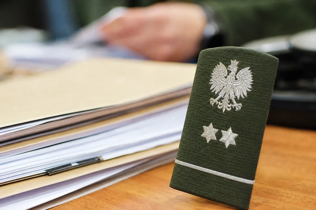 Zielony polski mundurowy naramiennik z orłem i dwiema gwiazdkami leży na biurku obok dokumentów, w tle dłonie osoby przeglądającej papiery.