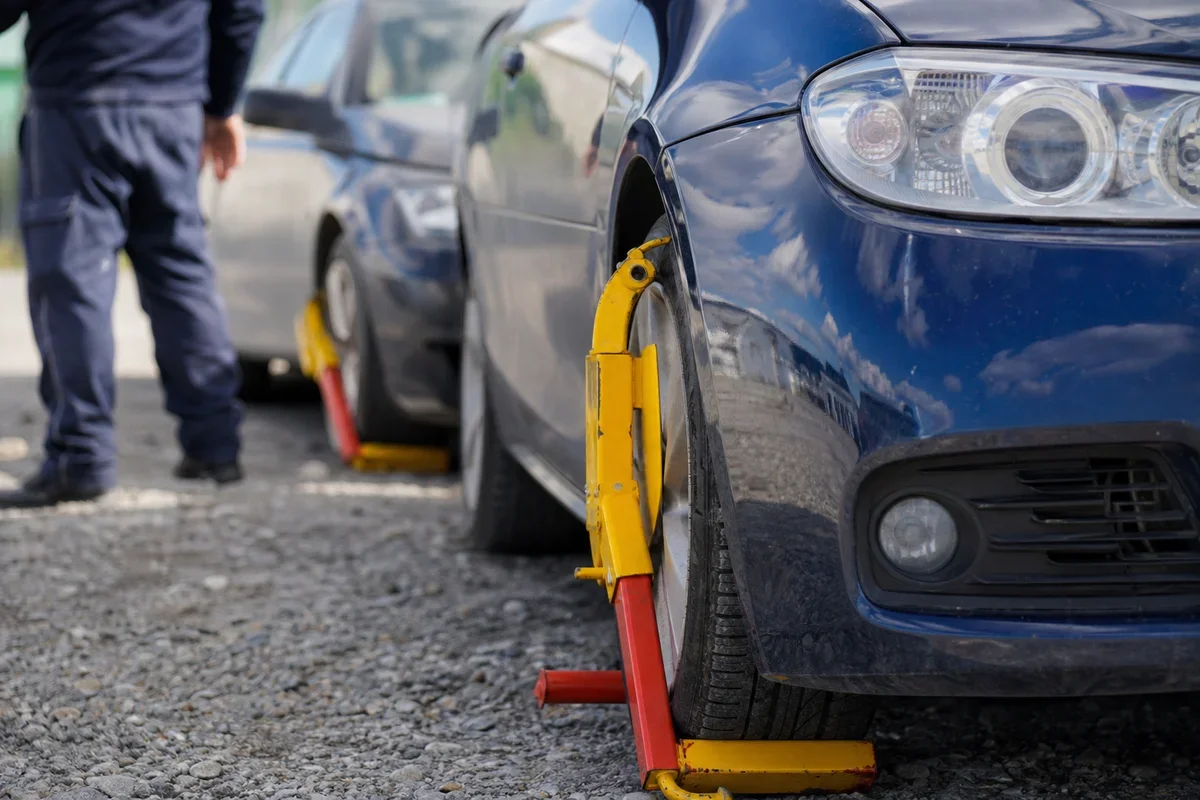 Samochody osobowe z założonymi blokadami na koła stoją na parkingu, w tle widoczny funkcjonariusz w mundurze.