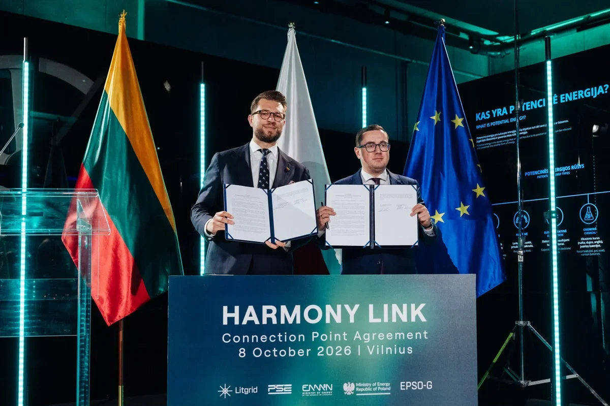 Dwaj mężczyźni w garniturach prezentują podpisane dokumenty podczas ceremonii Harmony Link w Wilnie, w tle flagi Litwy, Polski i UE.