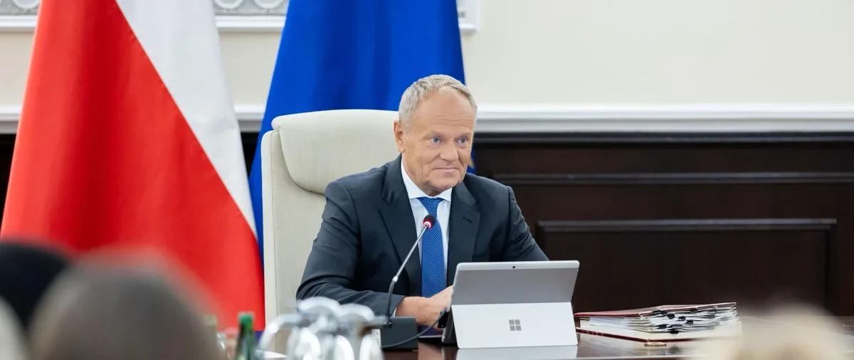 Mężczyzna w garniturze siedzi przy stole konferencyjnym z tabletem, przed nim mikrofon, w tle flagi Polski i Unii Europejskiej.