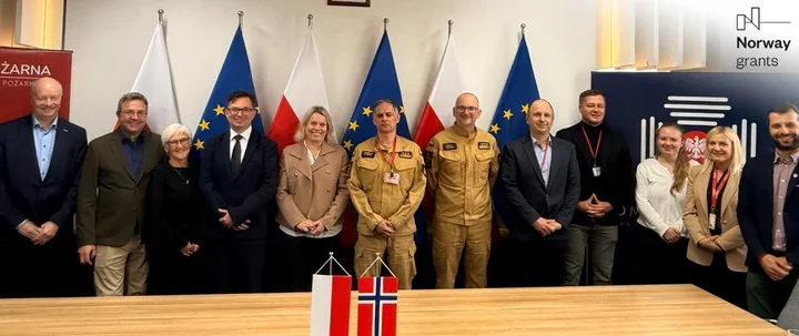 Grupa osób, w tym przedstawiciele służb mundurowych, stoi przy stole z flagami Polski i Norwegii podczas spotkania w siedzibie instytucji objętej wsparciem Norw