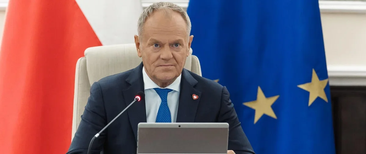 Mężczyzna w garniturze siedzi przy biurku z laptopem na tle flagi Polski i Unii Europejskiej.