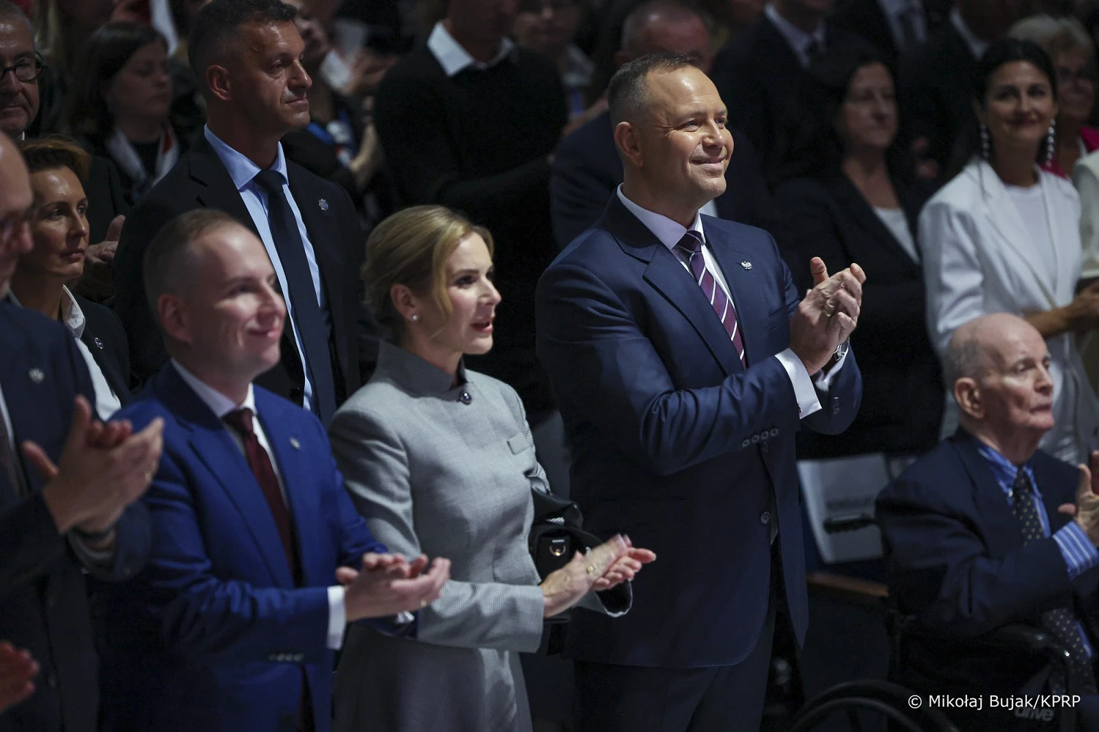 Grupa osób w eleganckich strojach stoi i klaska podczas oficjalnej uroczystości w sali widowiskowej.