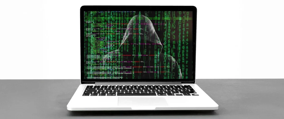 Laptop na szarym blacie, na ekranie widoczny sylwetka osoby w kapturze na tle zielonego kodu przypominającego efekt z filmu Matrix.