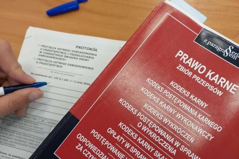 Ręka osoby wypełniającej protokół przy czerwonym zbiorze przepisów prawa karnego leży na drewnianym biurku.