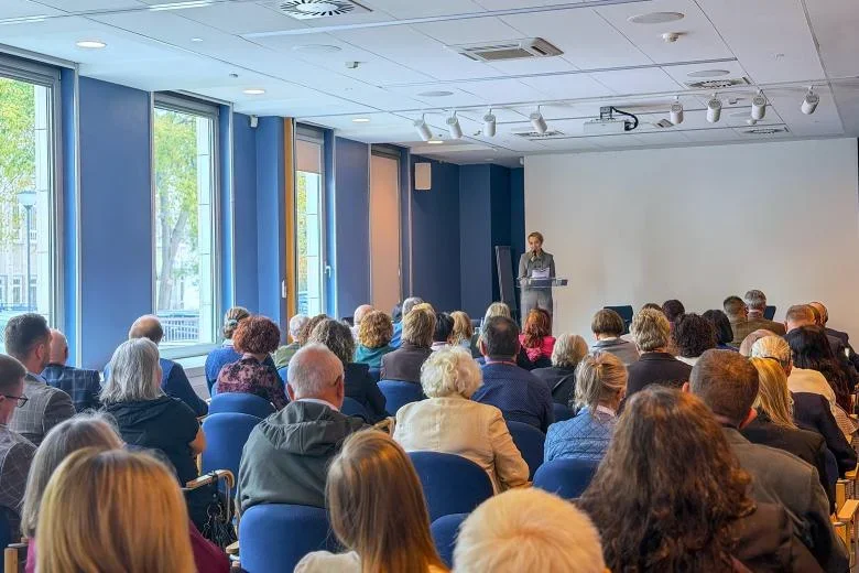 Mężczyzna wygłasza przemówienie zza pulpitu przed siedzącą publicznością w nowoczesnej sali konferencyjnej z dużymi oknami.