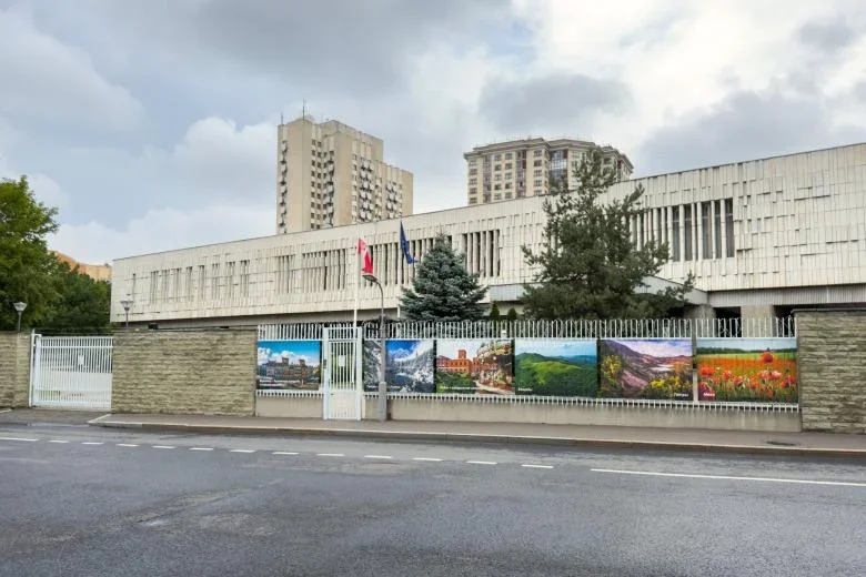 Budynek ambasady za kamiennym ogrodzeniem z plakatami krajobrazowymi, przed nim ulica i flagi.