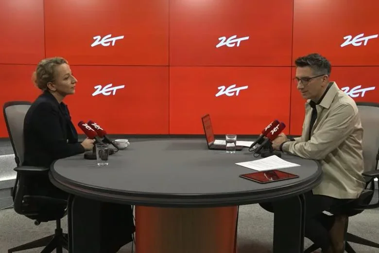 Dwie osoby siedzą przy okrągłym stole w studiu radiowym z czerwonym tłem z logo ZET, przed sobami mają mikrofony z logo ZET.