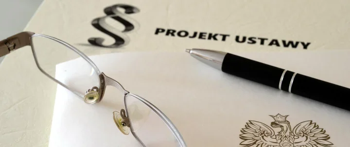 Na biurku leży dokument oznaczony napisem „PROJEKT USTAWY” z polskim orłem, długopis i okulary.