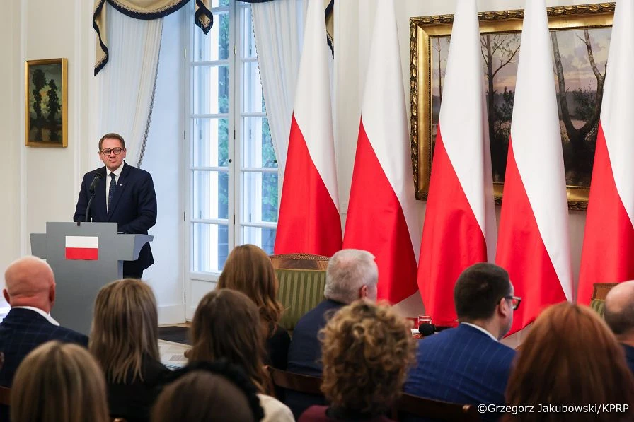 Mężczyzna w garniturze przemawia z pulpitu z flagą polską do publiczności w reprezentacyjnym pomieszczeniu z flagami Polski w tle.