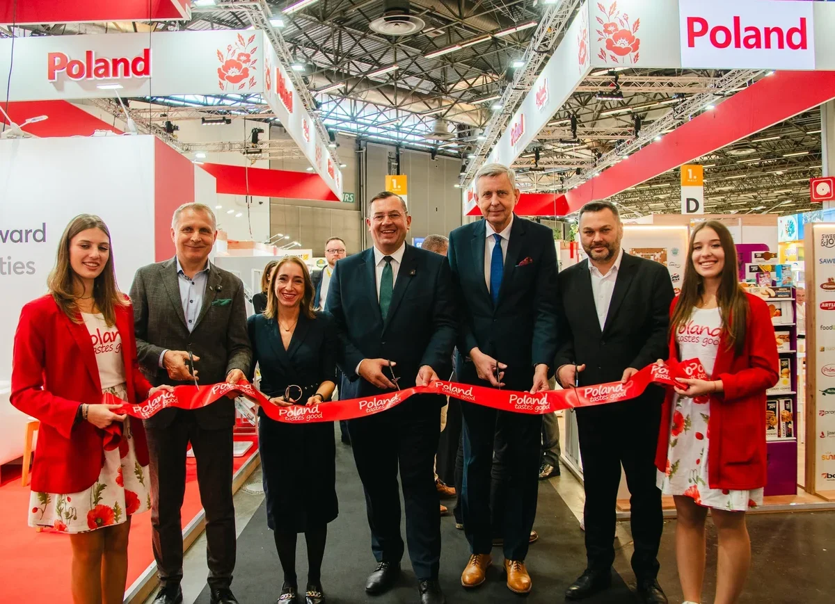 Grupa osób przecina czerwoną wstęgę na stoisku Polski podczas targów branżowych w hali wystawienniczej.