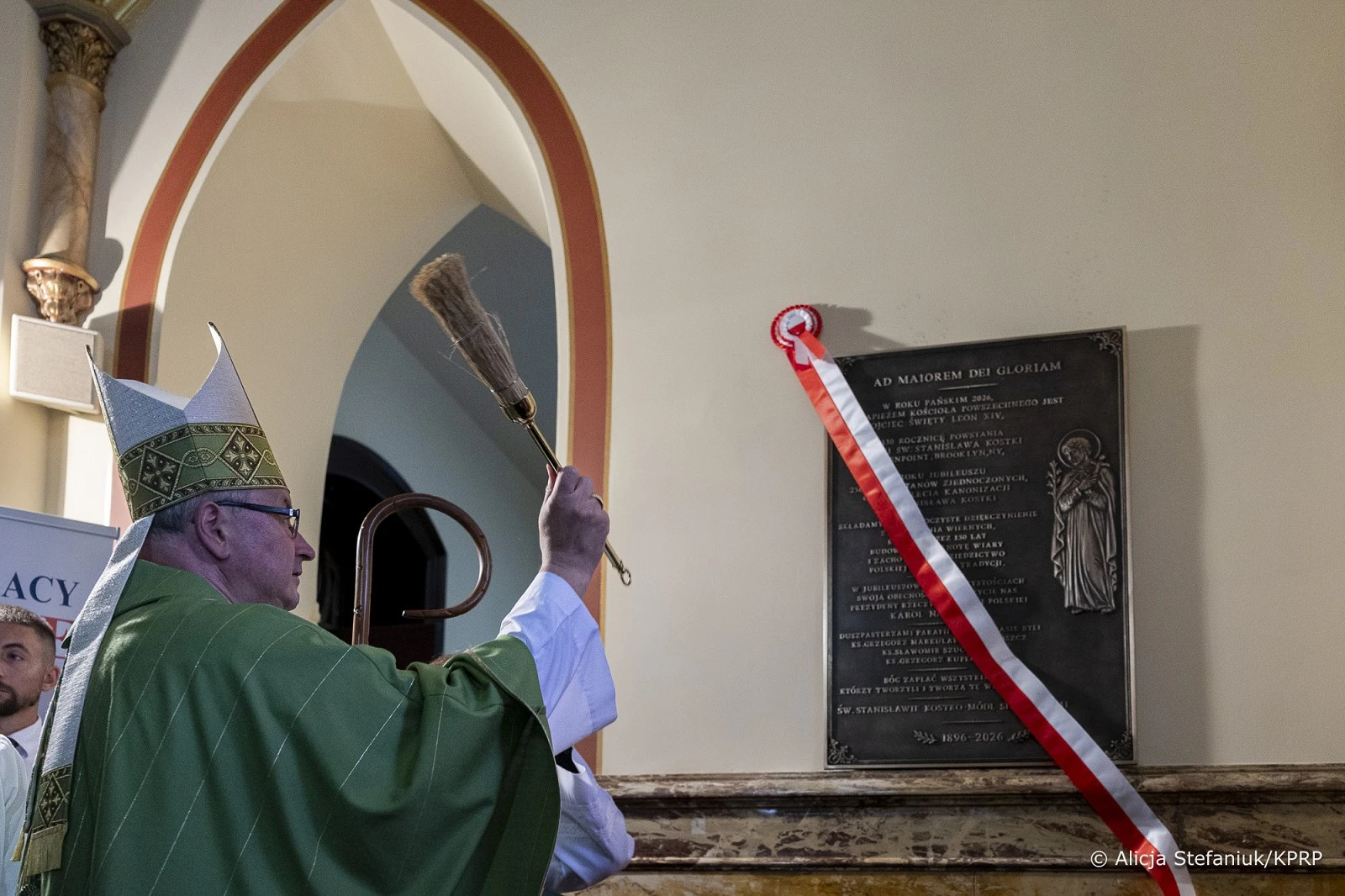 Biskup w zielonej szacie liturgicznej i mitrze poświęca wodą święconą tablicę pamiątkową w kościele.