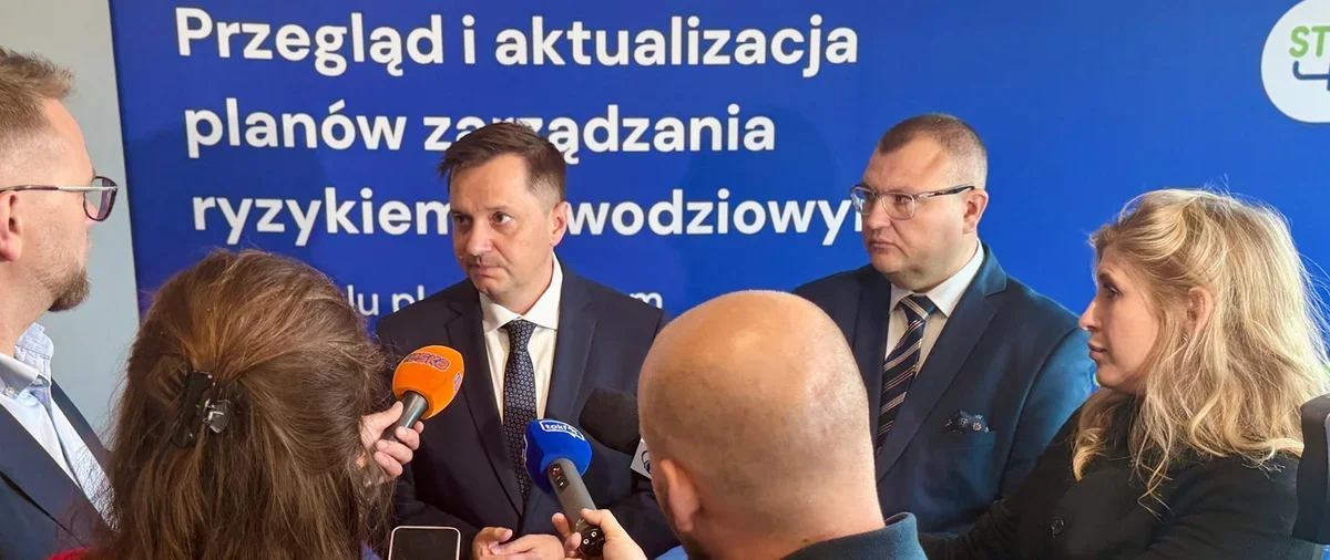 Trójka mówców w garniturach udziela wywiadu dziennikarzom podczas konferencji o zarządzaniu ryzykiem powodziowym.