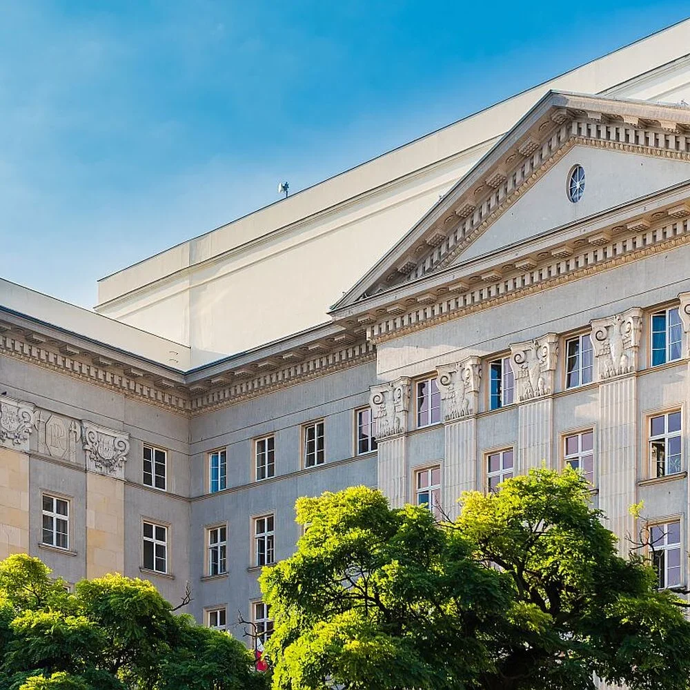 Fragment klasycystycznego budynku z kolumnami i tympanonem, częściowo zasłoniętego zielonymi drzewami, na tle błękitnego nieba.