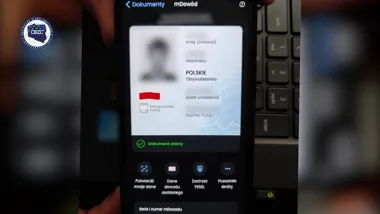 Ręka trzyma smartfon wyświetlający aplikację mDowód z elektronicznym dowodem osobistym, w tle klawiatura laptopa.