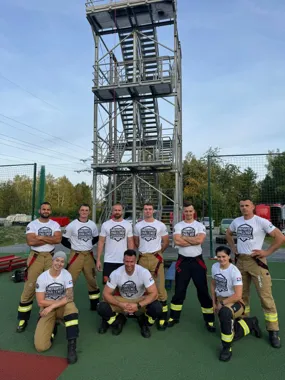 Grupa strażaków w koszulkach Firefighter Challenge pozuje do zdjęcia na boisku przed wieżą treningową.