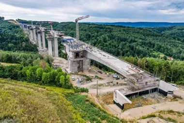 Budowa wielkiego mostu wiaduktu nad zalesioną doliną, z żurawiami, betonowymi filarami i częściowo ukończonym przęsłem mostu.