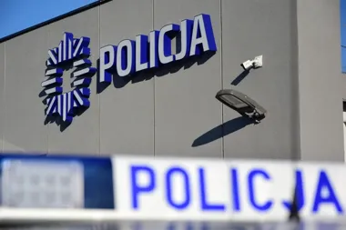 Fragment elewacji budynku z napisem i emblematem polskiej policji oraz policyjna taśma ostrzegawcza na pierwszym planie.